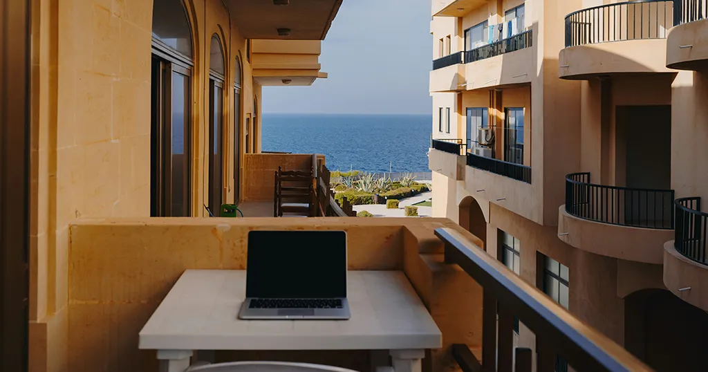 A laptop open on a sunlit balcony above the Mediterranean, the sea filling the gap between the buildings — the frame is at home, you are not