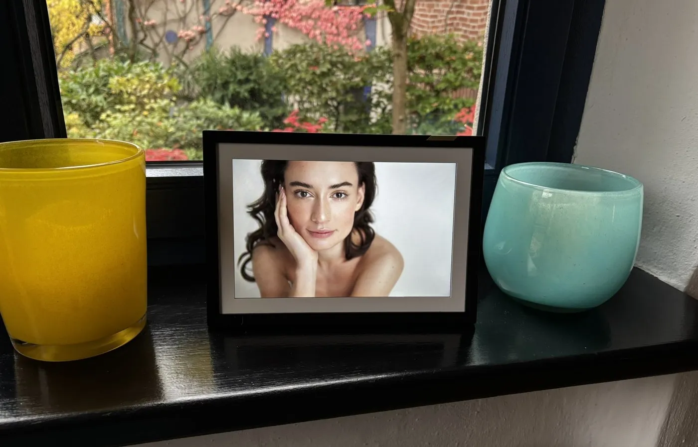 The Nixplay Classic 10.1-inch touch screen photo frame on a windowsill between a yellow and a turquoise glass vase